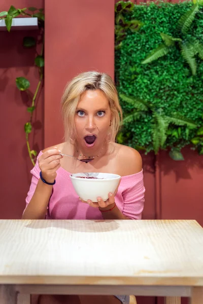 Woman looking shocked to camera while eating an acai smoothie bowl in a cafe. Healthy eating concept.