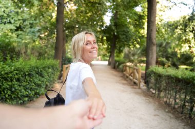 Genç bir kadın kameraya bakıyor ve el ele tutuşurken gülümsüyor ve birini parkta yürüyüşe çıkarıyor..