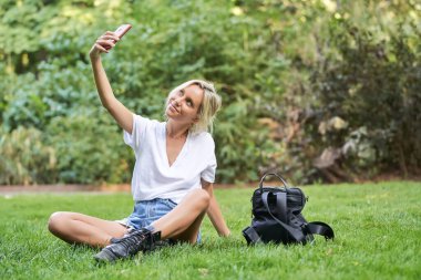 Dışarıda çimlerin üzerinde otururken cep telefonuyla selfie çeken genç bir kadın. Teknoloji konsepti.