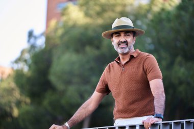 Şapkalı orta yaşlı bir adam kameraya bakıyor ve dışarıda korkuluklara yaslanırken gülümsüyor..