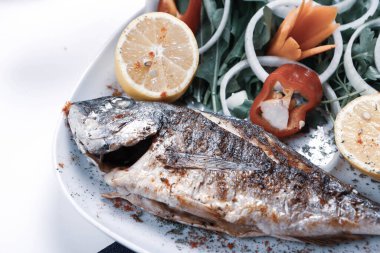 Kızarmış deniz çerezi (cupra) beyaz tabakta soğan, bahçe roketi ve limon dilimli balık.