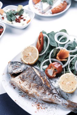 Kızarmış deniz çerezi (cupra) beyaz tabakta soğan, bahçe roketi ve limon dilimli balık.