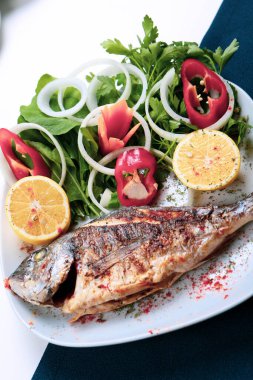 Kızarmış deniz çerezi (cupra) beyaz tabakta soğan, bahçe roketi ve limon dilimli balık.