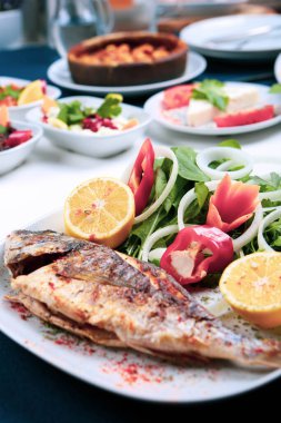 Kızarmış deniz çerezi (cupra) beyaz tabakta soğan, bahçe roketi ve limon dilimli balık.