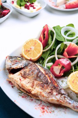 Kızarmış deniz çerezi (cupra) beyaz tabakta soğan, bahçe roketi ve limon dilimli balık.
