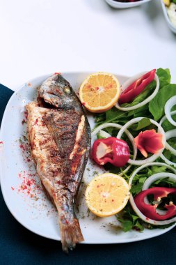 Kızarmış deniz çerezi (cupra) beyaz tabakta soğan, bahçe roketi ve limon dilimli balık.