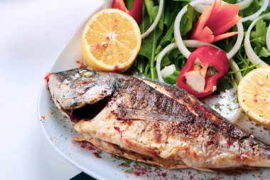 Kızarmış deniz çerezi (cupra) beyaz tabakta soğan, bahçe roketi ve limon dilimli balık.