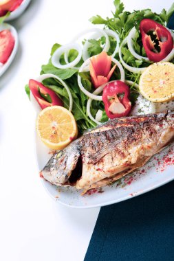 Kızarmış deniz çerezi (cupra) beyaz tabakta soğan, bahçe roketi ve limon dilimli balık.