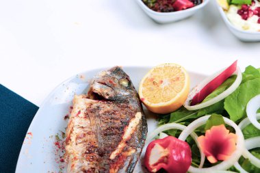 Kızarmış deniz çerezi (cupra) beyaz tabakta soğan, bahçe roketi ve limon dilimli balık.