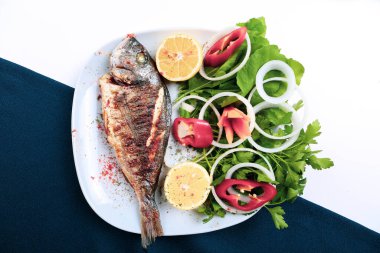 Kızarmış deniz çerezi (cupra) beyaz tabakta soğan, bahçe roketi ve limon dilimli balık.