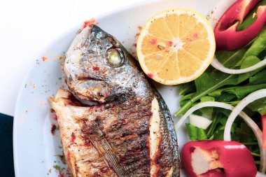 Kızarmış deniz çerezi (cupra) beyaz tabakta soğan, bahçe roketi ve limon dilimli balık.