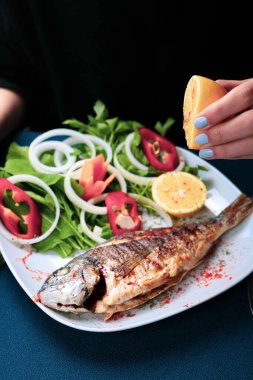 Kızarmış deniz çerezi (cupra) beyaz tabakta soğan, bahçe roketi ve limon dilimli balık.