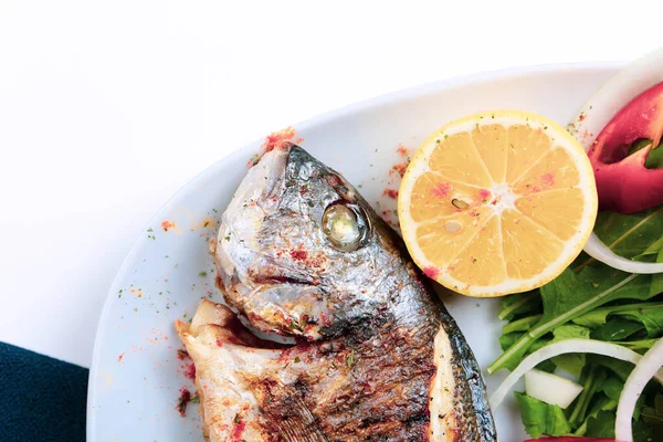 Kızarmış deniz çerezi (cupra) beyaz tabakta soğan, bahçe roketi ve limon dilimli balık.