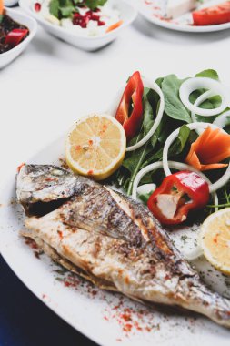 Kızarmış deniz çerezi (cupra) beyaz tabakta soğan, bahçe roketi ve limon dilimli balık.