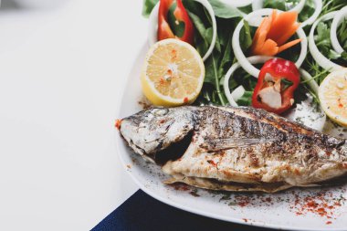 Kızarmış deniz çerezi (cupra) beyaz tabakta soğan, bahçe roketi ve limon dilimli balık.