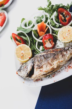 Kızarmış deniz çerezi (cupra) beyaz tabakta soğan, bahçe roketi ve limon dilimli balık.