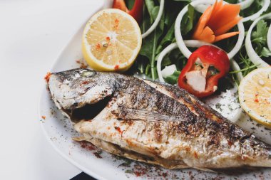 Kızarmış deniz çerezi (cupra) beyaz tabakta soğan, bahçe roketi ve limon dilimli balık.