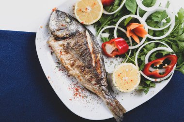 Kızarmış deniz çerezi (cupra) beyaz tabakta soğan, bahçe roketi ve limon dilimli balık.