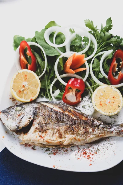 Kızarmış deniz çerezi (cupra) beyaz tabakta soğan, bahçe roketi ve limon dilimli balık.