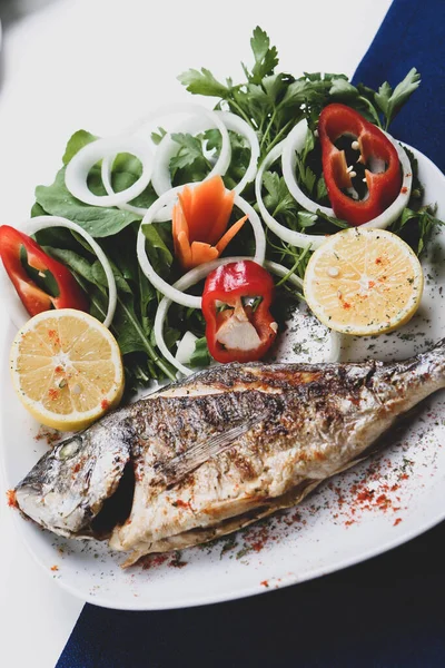 Kızarmış deniz çerezi (cupra) beyaz tabakta soğan, bahçe roketi ve limon dilimli balık.