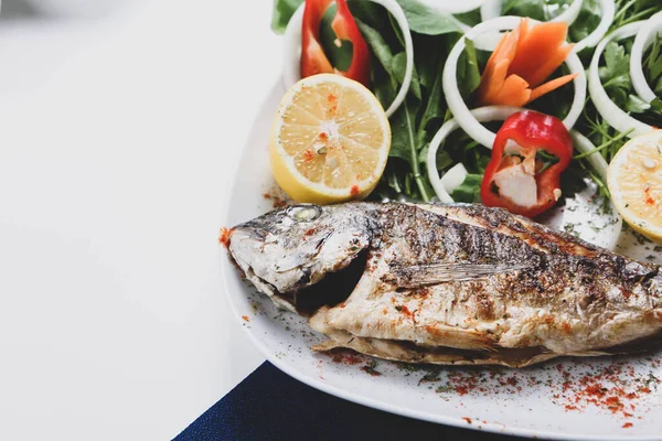 Kızarmış deniz çerezi (cupra) beyaz tabakta soğan, bahçe roketi ve limon dilimli balık.