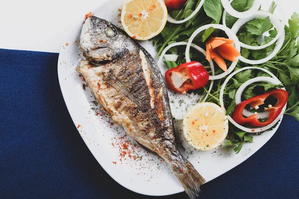 Kızarmış deniz çerezi (cupra) beyaz tabakta soğan, bahçe roketi ve limon dilimli balık.