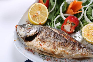 Kızarmış deniz çerezi (cupra) beyaz tabakta soğan, bahçe roketi ve limon dilimli balık.