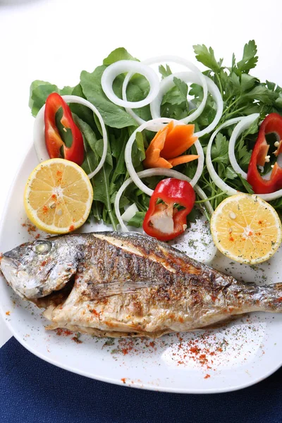 Kızarmış deniz çerezi (cupra) beyaz tabakta soğan, bahçe roketi ve limon dilimli balık.