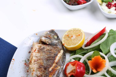 Kızarmış deniz çerezi (cupra) beyaz tabakta soğan, bahçe roketi ve limon dilimli balık.