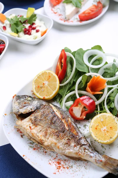 Kızarmış deniz çerezi (cupra) beyaz tabakta soğan, bahçe roketi ve limon dilimli balık.