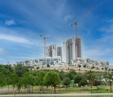 Modiin, İsrail, 21 Mayıs 2022. Çok katlı bir binanın inşaatı. Ağaçlı yeşil bir park ve konut binalarının yanında beyaz bulutlu mavi bir gökyüzüne karşı bir çimenlik.