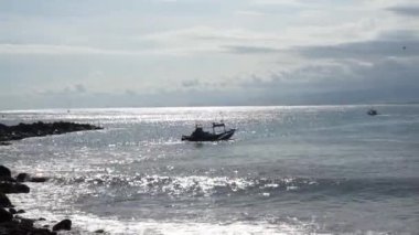 Güneşin deniz suyundaki yansımasıyla güzel bir sahil manzarası var. Balıkçı teknelerinin geçişini izliyor.