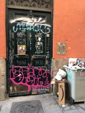 Madrid, İspanya; 02 24 2019. Madrid 'in merkezindeki bir sokakta kapı grafiti ile tahrip edilmiş. Kirli ve kirli bir binanın kapısı taşan bir çöp konteynerinin yanında..