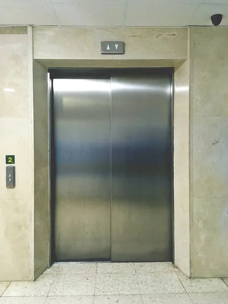 Door of a closed elevator with the buttons to call it to the side. Metal door in a marble wall. Elevator installed on floor 2 of a hospital in Cordoba, Spain.