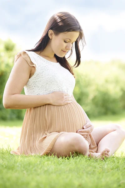 Gravid kvinna sitter på gräset utomhus håller bump — Stockfoto