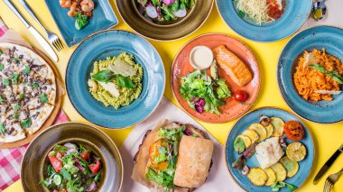 Izgarada somon bifteği, biftekli morina, etli burger, karidesli fusili, mantarlı pizza, beyaz ahşap masada ızgara biftekli salata.