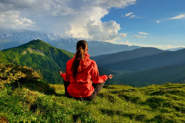 Молода жінка в червоній куртці сидить в позі йоги в горах — стокове фото