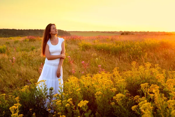 Jovem mulher de roupas brancas em pé no campo ao pôr do sol — Fotografia de Stock