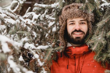Kış şapkası takmış şık sakallı bir adam soğuk havada kozalaklı bir ormanda duruyor ve güzelce gülümsüyor. Buz mevsimi. Boşluğu kopyala