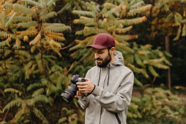 Elinde fotoğraf makinesi olan şık sakallı bir fotoğrafçı sonbahar parkında fotoğraf çekiyor.