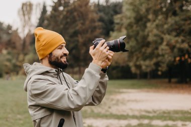Elinde fotoğraf makinesi olan şık sakallı bir fotoğrafçı sonbahar parkında fotoğraf çekiyor.