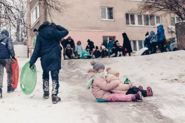 23.11.2018 Vinnitsa, Ukrayna: küçük kız, çocukların kış eğlencesini geçirdikleri şehirdeki yüksek binaların bahçesinde kendinden büyük çocukların yanında buz kayağına biniyor.