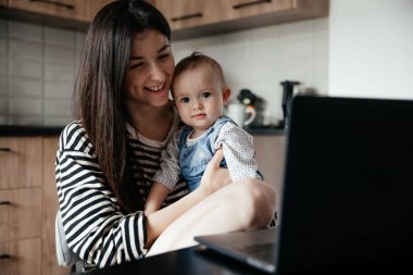 Bir sürü çocuğu olan genç bir anne mutfakta kucağında bir bebekle otururken büyük kızı mutfakta yemek ve temizliğe yardım ediyor.