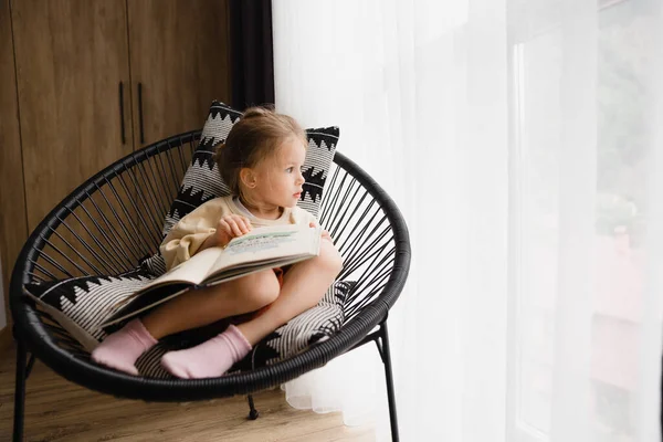 Μικρό Κορίτσι Κάθεται Μια Καρέκλα Δίπλα Στο Παράθυρο Διαβάζοντας Ένα — Φωτογραφία Αρχείου