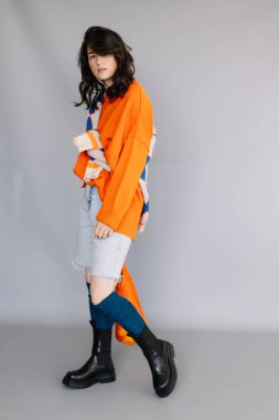 Turuncu örgü ceket giymiş şık bir kadın gri bir arka planda fotoğraf stüdyosunda fotoğraf çektirmek için poz veriyor. Parlak duygular, pozlar, modern konsept