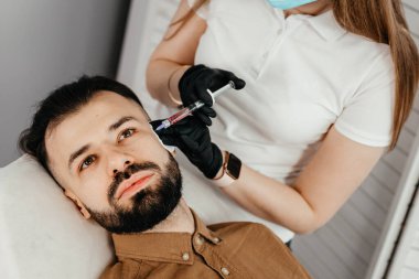 Stylish bearded man visiting aesthetic clinic, getting lips filler, closeup. Man having beauty injection at male spa salon. Anti-aging treatment for men concept