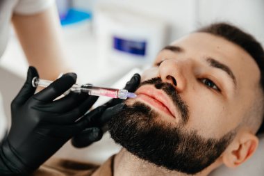 Stylish bearded man visiting aesthetic clinic, getting lips filler, closeup. Man having beauty injection at male spa salon. Anti-aging treatment for men concept