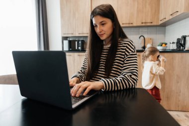 Serbest çalışan kadın, mutfak ofisindeki masanın başında oturup dizüstü bilgisayarla çalışıyor. Oyuncu çocuk dikkatini işten uzaklaştırıyor, çocuk gürültü yapıyor ve meşgul annesinden ilgi istiyor.