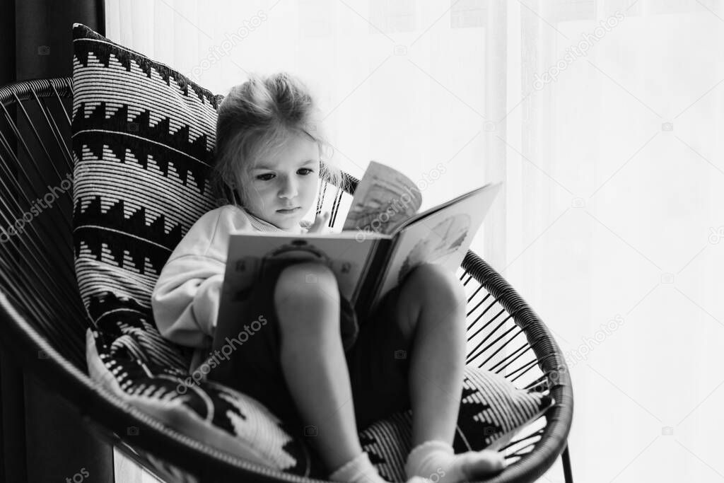 child looks at the pages of a book sitting in a comfortable chair on the balcony of the apartment during the day