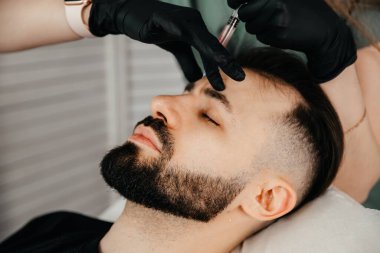Stylish bearded man visiting aesthetic clinic, getting lips filler, closeup. Man having beauty injection at male spa salon. Anti-aging treatment for men concept